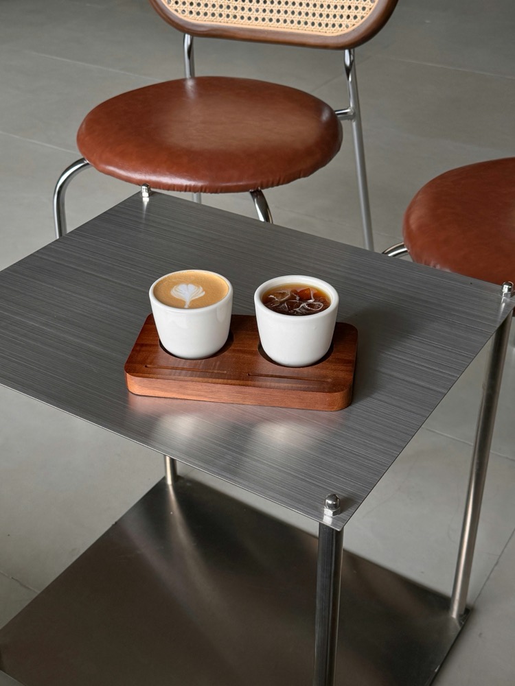 Zwei Kaffeetassen auf einem Holzbrett, Tisch aus gebürstetem Stahl, Stühle mit Lederpolster